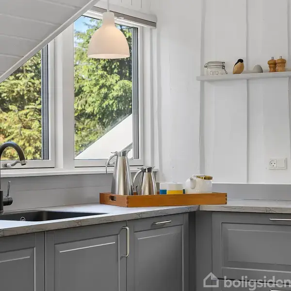 Et køkkenhjørne med grå skabe og en sort vandhane ved siden af et vindue. En bakke med ståltermokander står på bordet, mens hylder har små dekorationer.
