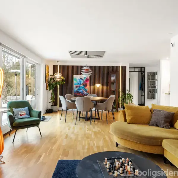 En stue med en stor gul sofa, et rundt sofabord med skakbrikker og en høj orange lampe. Spiseområde med rundt bord og stole, omgivet af kunst og store vinduer.