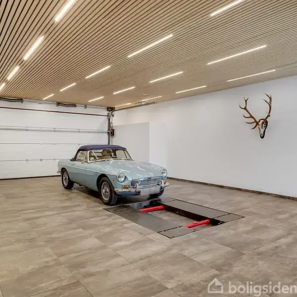 Blå vintage cabriolet holder stille i en stor, moderne garage med flisegulv. Loftet har træpaneler, og et hjortegevir hænger på væggen.