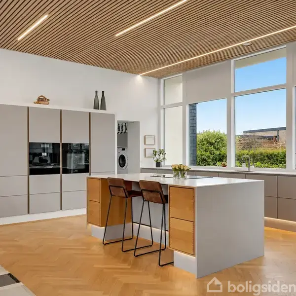 Moderne terrasse med spisebordssæt i træ, omgivet af frodig hæk. Store vinduer giver udsigt til husets indre. Blå himmel og nabohus ses i baggrunden.