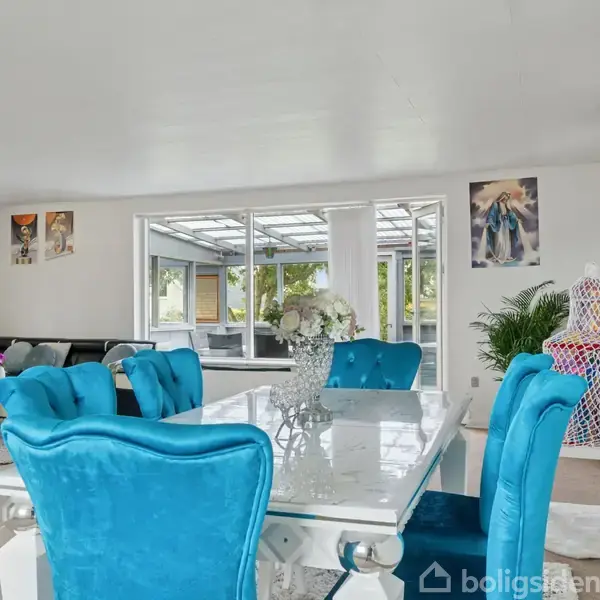 Spisebord med blå polstrede stole står i et lyst rum. Bordet er pyntet med en blomsterdekoration. I baggrunden ses en sofagruppe foran store vinduer med udsigt til terrasse.