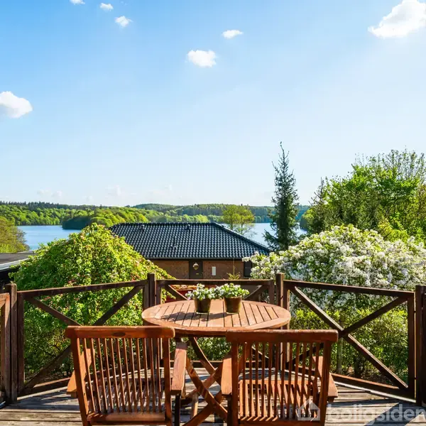 Et rundt træbord med fire stole står på en terrasse. Bordet har en række planter. I baggrunden ses frodig vegetation, en sø, og en blå himmel med spredte skyer.