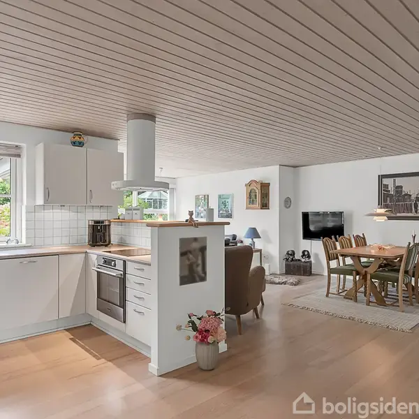Køkkenet er lyst og moderne med hvidt inventar og integrerede apparater. Ved siden af sidder en åben stue, som har spisebord og opholdsområde. Store vinduer giver udsigt til haven.