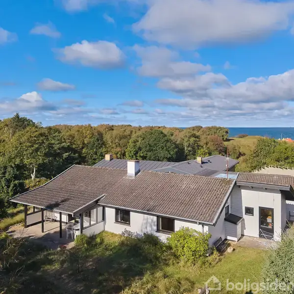 Hus omgivet af grønne træer, ses fra oven. Det har en gråbrun tag og hvid facade. I baggrunden anes havet under en blå himmel med skyer.