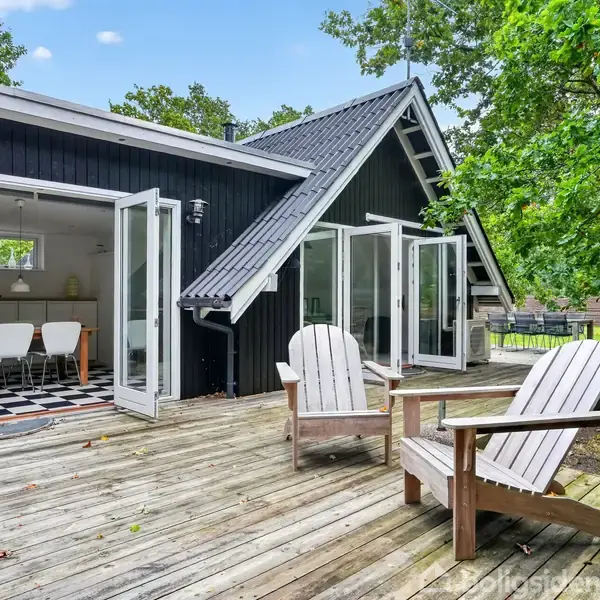 En sortmalet træhytte står med åbne, hvide dobbeltdøre til en terrasse, hvor to træstole er placeret. Indenfor ses spisebordstole, og omgivelserne er grønne træer.