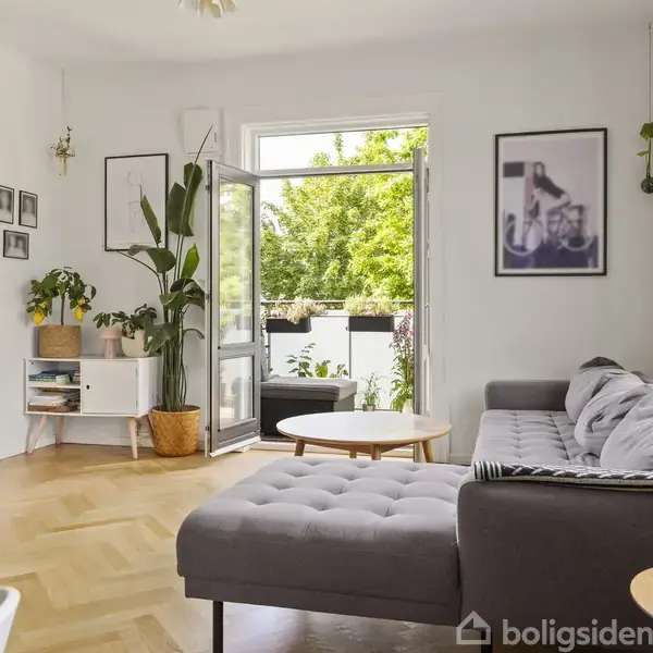 Grå sofa står i en lys stue med trægulv, hvor døren til en altan står åben. Væggene har indrammede billeder, og der er planter i hjørnerne.