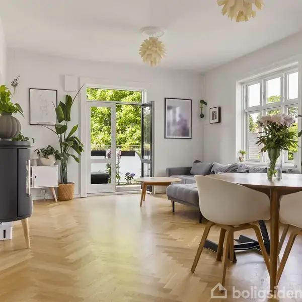 Et lyst stue-miljø med parketgulv og moderne møbler. Et rundt spisebord med blomster i vase, sofaer og store planter. Store vinduer giver udsigt til grønne træer.