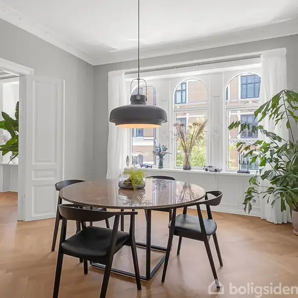 Rundt spisebord med sorte stole står i et lyst rum med store vinduer. En hængelampe hænger over bordet. Grønne planter står rundt omkring. En tv-stue er synlig i baggrunden.