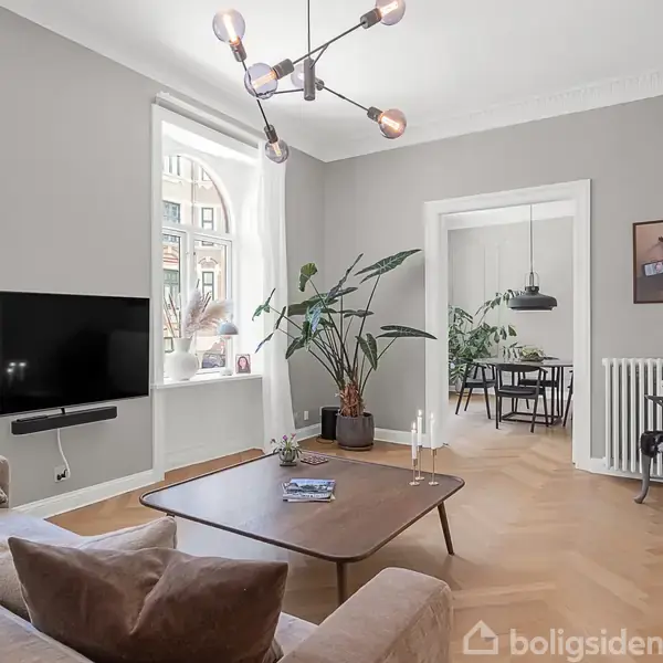 Stue med grå vægge, en sofa, fladskærms-TV på væggen og et sofabord. En stor plante står ved vinduet. I baggrunden ses en spisestue med bord og lamper.