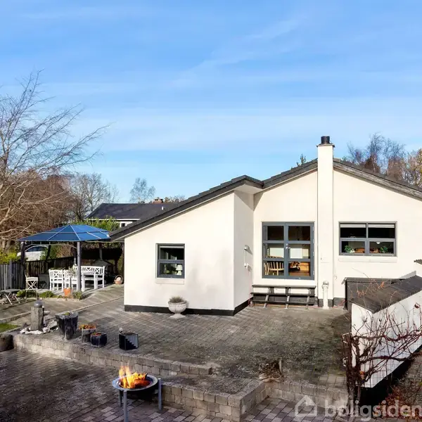 Hvid hus med skråtag står på en flisebelagt terrasse. En åben pejs er tændt. Til venstre står et havebord under en blå pavillon. Omkring er der træer og græsplæne.
