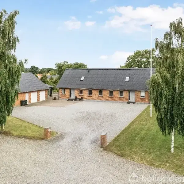 En rød murstensbygning med sort tag står omkranset af grusplads og græsplæner. To høje træer i forgrunden, med en flagstang til højre. Klar, blå himmel.