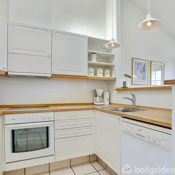 Et hvidt køkken med træbordplader indeholder en ovn, opvaskemaskine og hylder med krukker. Rummet er oplyst af hængelamper, og væggene har indrammede billeder.