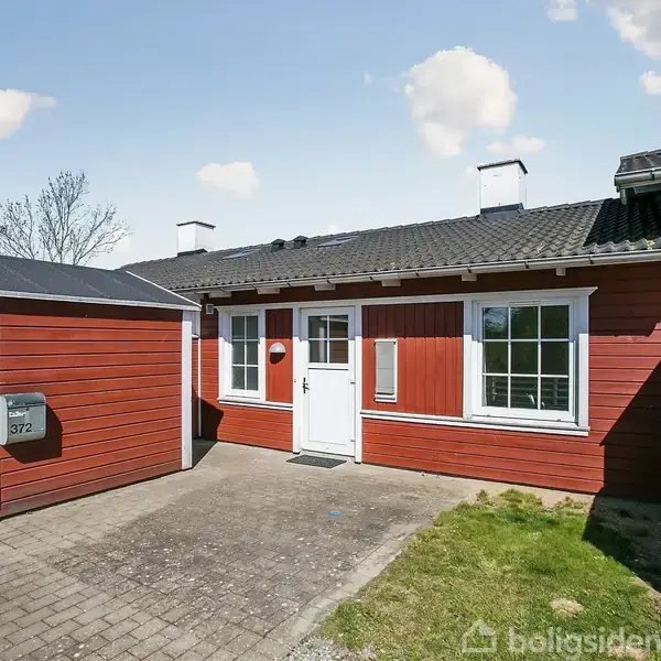 Rødt rækkehus med hvide vinduer og dør, står ved siden af et skur. Beliggende i en solrig have med grøn græsplæne. Husnummeret er 372.