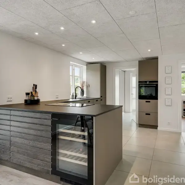 Et moderne køkken med en stor køkkenø, der har indbygget vinkøleskab. Køkkenet er lyst med cremefliser og indbyggede loftspots. I baggrunden ses spiseplads og dør til terrasse.