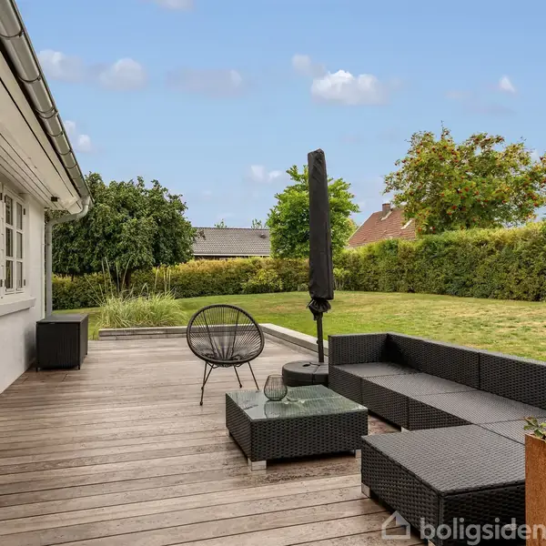 Terrassemøbler står på en træterrasse ved siden af et hus. Sofaen og et bord er placeret bag en foldet parasol. Baggrunden viser en have med græs og hække.