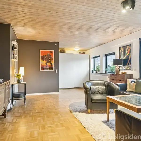 Stue med mørk lædersofa omkring et sofabord; plakater på vægge, døre til højre, og vinduer med blå gardiner. Parquetgulv og træloft skaber en hyggelig atmosfære.