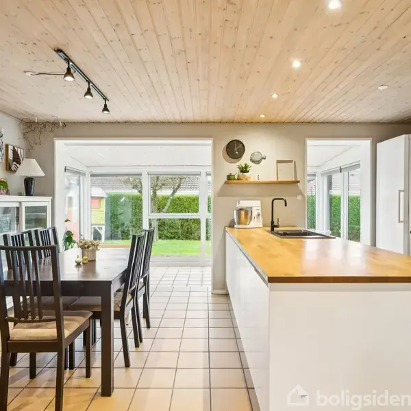 Et lyst køkken med et træloft har en køkkenø med vask. Et spisebord med fire stole står ved siden af. Store vinduer viser en grøn have udenfor.