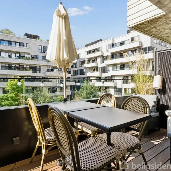 Terrassemøbler med parasol står på træterrasse. Fire stole om et bord med udsigt til moderne lejlighedskompleks i baggrunden. Grønne træer omgiver området.