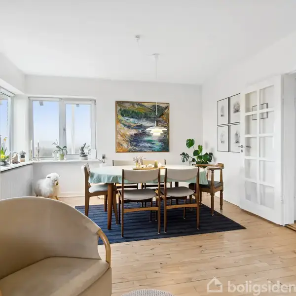 Stue med spisebord står på sort tæppe, omkranset af seks stole. Væggene er hvide med kunst og glasdør. Lyst parketgulv og grønne planter fuldender rummet.