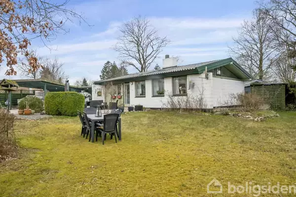 En lav, hvid bungalow med grønne tagkanter står i en stor have med græsplæne. Et sort udendørs spisebord med seks stole er placeret på plænen.