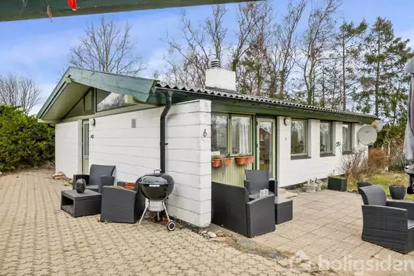 Et enkelt sommerhus med hvid træbeklædning står stille på en flisebelagt terrasse. Sorte havemøbler og en grill er placeret udenfor. Omgivet af høje træer i baggrunden.