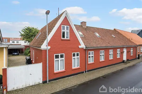 Rødt murstenshus med hvide vinduesrammer står stille på en gade. Ved siden af husets port ses en overdækket indkørsel med en bil. Huset har et rødt tegltag.