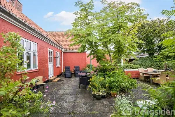 Rødt murstenshus med hvide vinduer, omkranset af frodig have. Terrasse med havemøbler og blomsterpotter. Et bord og stole står under et stort grønt træ.