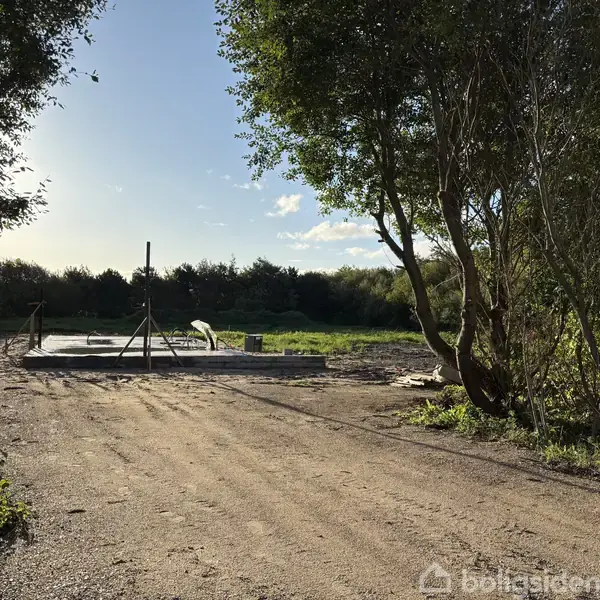 Byggeplads under opførelse med fundament og arbejdsredskaber, omgivet af en grusvej og træer. Vejret er solrigt med klar himmel og grønne omgivelser.