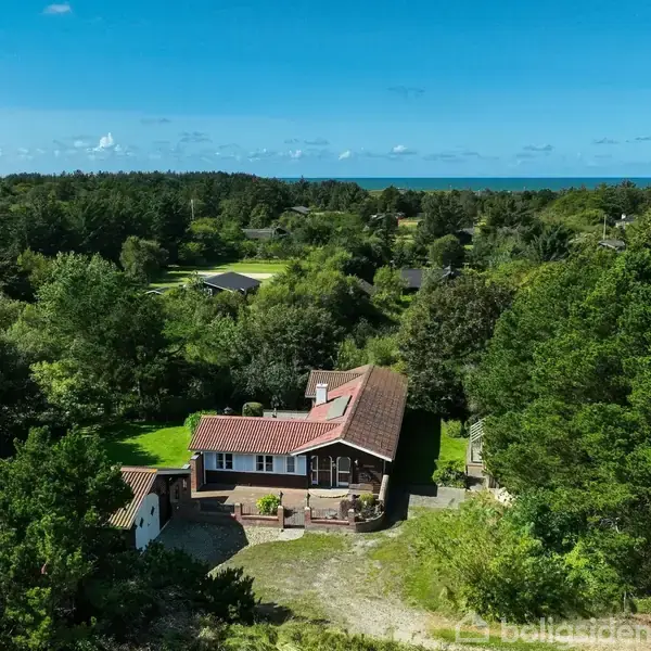 Et rødt taghus står stille omgivet af frodig skov. I baggrunden ses et stort grønt landskab og havet i horisonten. Bright sollys oplyser scenen.