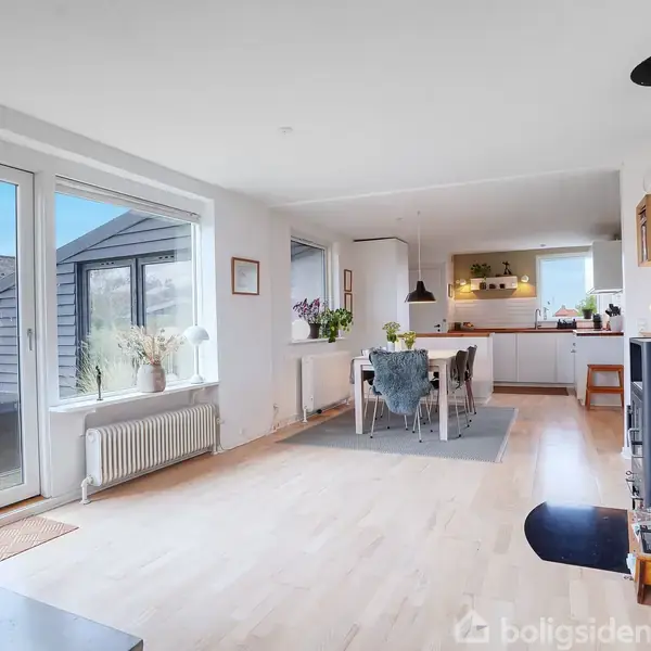 Stue med brændeovn til højre, spisebord i midten. Køkken i baggrunden. Lyse trægulve, store vinduer og en terrassedør til venstre. Indbyder til lys og rummelighed.