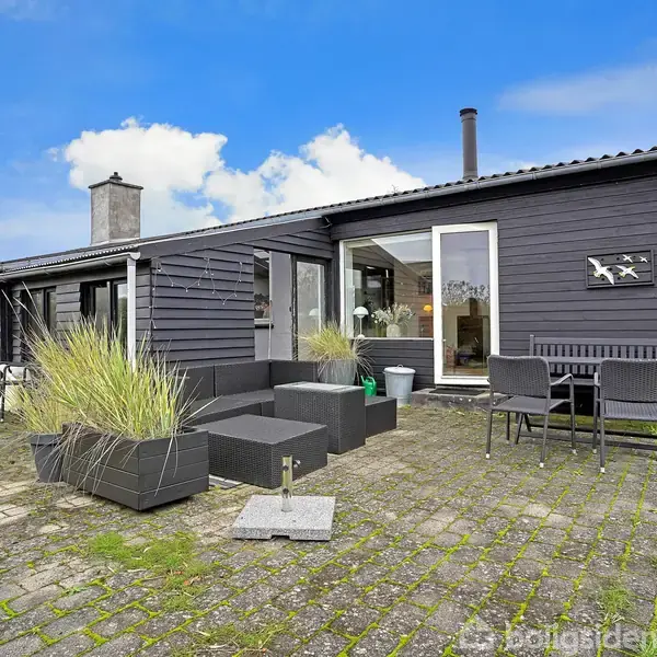 Et sortmalet hus med store vinduer står på en stenterrasse med havemøbler og plantekasser. Terrassen er omgivet af grønne planter og under en blå himmel med hvide skyer.