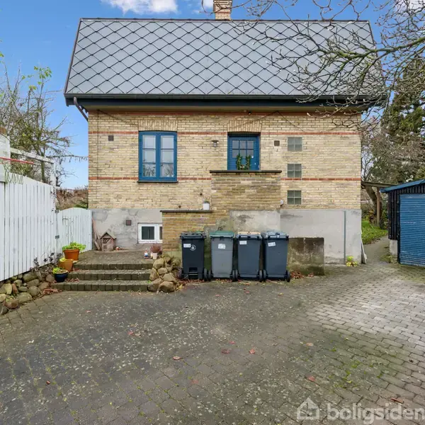 Hus med gult murværk står stille i en gårdhave omgivet af grønne træer. Foran huset er der tre affaldscontainere vendt mod en blå garage. En hvid træhegn flankerer en stentrappe.