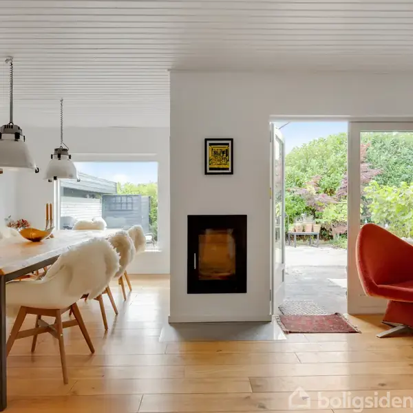 Et moderne spisebord med hvide stole står i en lys spisestue. To lamper hænger over bordet. En pejs er indbygget i væggen. Dør åbner ud til terrasse med have.