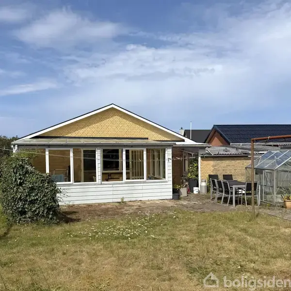 Et hus med gul murstensfacade og en udestue står i haven. Til højre ses et drivhus og havemøbler. Skyfri blå himmel i baggrunden.