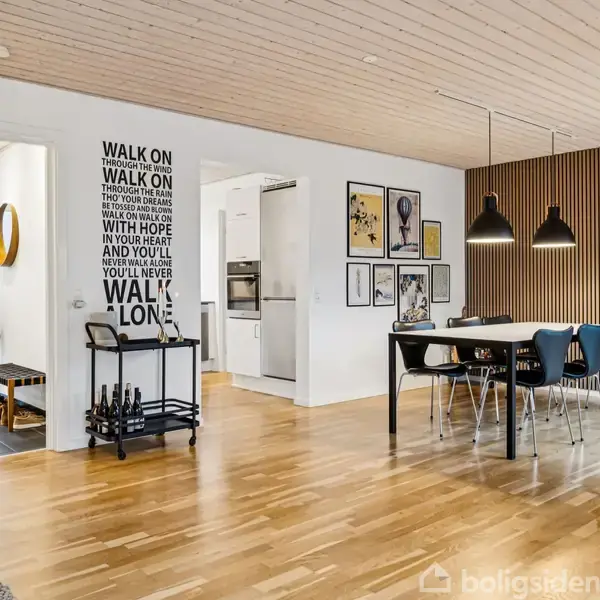 En moderne spisestue, hvor et sort bord og stole står under tre loftslamper. På væggen hænger der kunst. Til venstre står en brændeholder og indgangen til køkkenet. På væggen står tekst: "WALK ON WALK ON WITH HOPE IN YOUR HEART AND YOU'LL NEVER WALK ALONE."