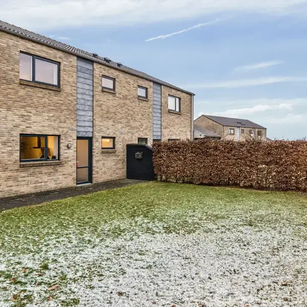 Et toetagers murstenshus står i en have med en snedækket græsplæne. Baggrunden viser flere lignende huse og en hæk omkring haven.