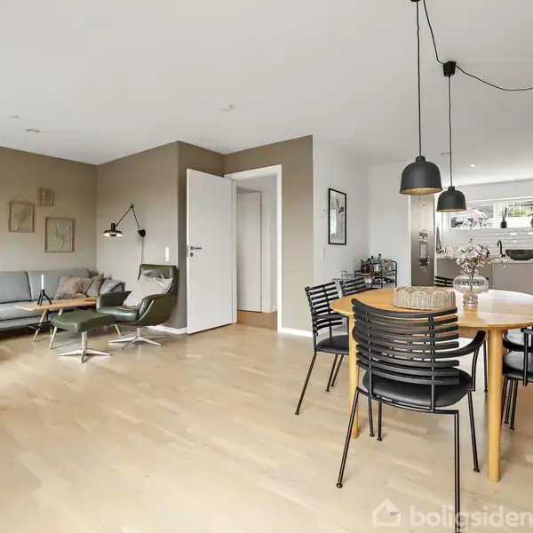 Stue-dining område med spisebord og sorte stole. Stuen har en grå sofa, lænestol, og små vægdekorationer. Rummet er lyst med trægulv, et åbent køkken til højre.
