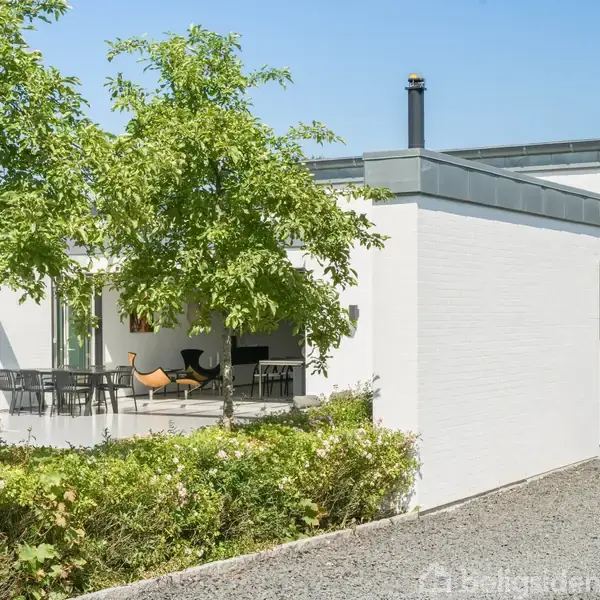 Hvid, moderne bygning står roligt. Terrasse med spisebord og stole ses under et grønt træ. Omkringliggende have har en grussti og buske i et solrigt miljø.