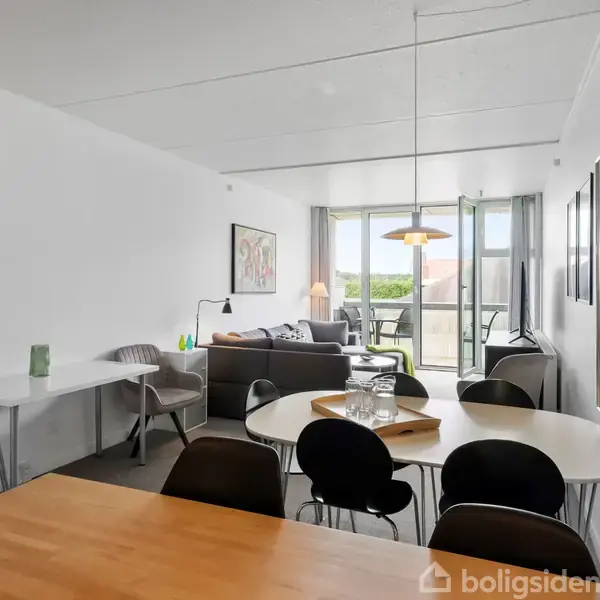 Et lyst, moderne stueområde med spisebord og stole i forgrunden. I baggrunden ses en sofa og glasdøre, der fører til en balkon. Plakat på væggen: "BLOKHUS".