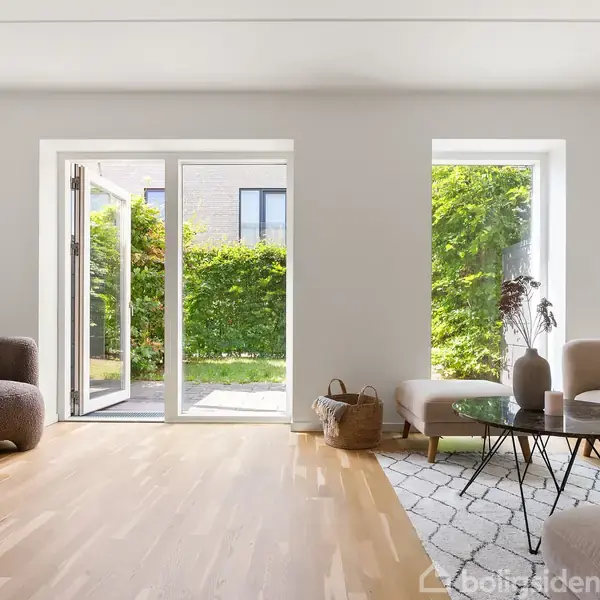 En lys stue med sofa og lænestol står over for glasdøre, der åbner ud til en grøn have. Rummet er dekoreret med et tæppe og et lavt bord.