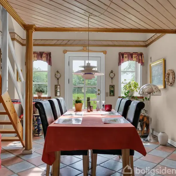 Et spisebord dækket med en rød dug, omgivet af stole, står i et rum med trægulv og en trappe til venstre. Glasdør og vinduer viser en grøn have udenfor.