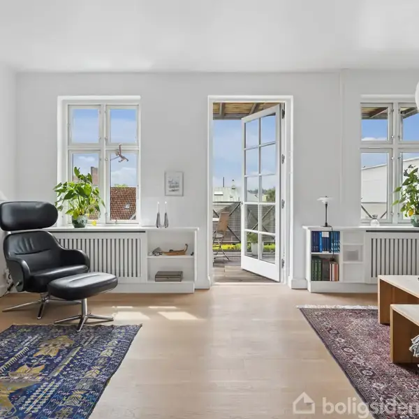 En lys stue med lænestol, sofabord og bogreol. Store vinduer og en åben dør fører ud til en altan. Indendørs planter ved vindueskarmen. Tæpper på trægulvet.