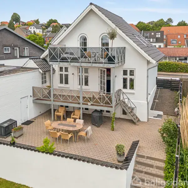 En balkon med kurvestole og et rundt bord med udsigt til en sø og en stor, historisk bygning i baggrunden. Solrig himmel med få skyer.