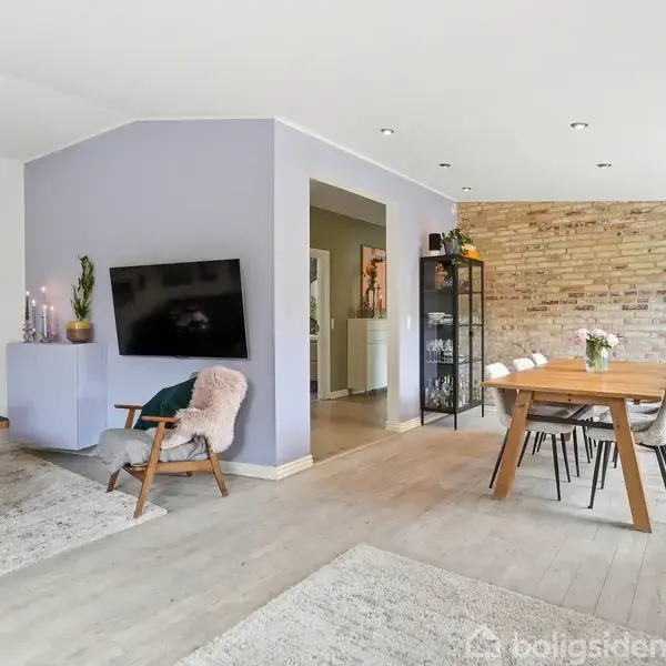 Stue med TV på væggen, træstol og bord med blomster; spisebord med stole. Lys træparketgulv, murede vægge og åbning til køkkenet skaber en lys og hyggelig atmosfære.