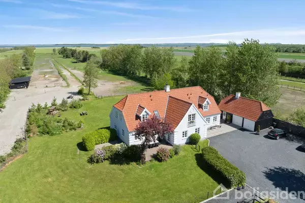 Hvidt hus med rødt tegltag står på en velplejet græsplæne omgivet af buske, med en grusvej og marker i baggrunden under en blå himmel.