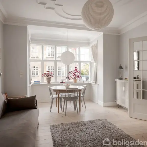 En stue med en grå sofa til venstre, et rundt spisebord med blomster på, og store vinduer bagved. En dør åbner til et tilstødende rum.
