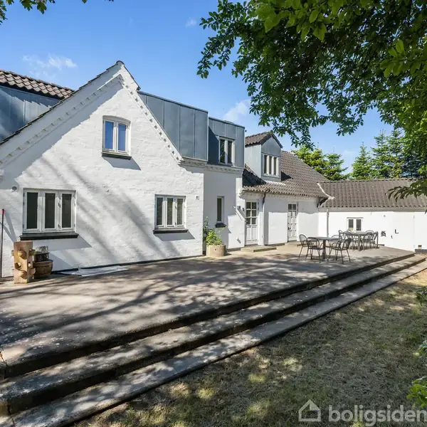 Et hvidt hus med sort tag står i sollys. En stor terrasse med trapper foran indeholder et bord og stolearrangement. Grønne træer omgiver stedet og giver skygge.