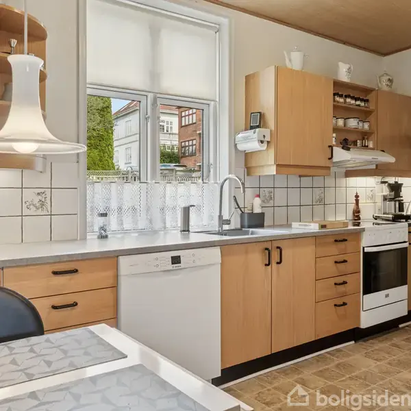 Køkkenet har træskabe, hvidt flisebelagt væg, og en kogeplade med ovn. En lampe hænger over et bord med dækkeservietter. Vinduerne viser en udsigt til et hus.