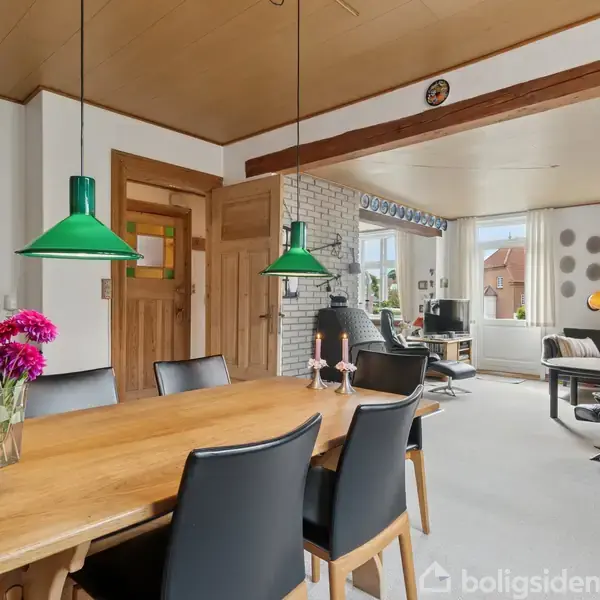 Et træbord med sorte stole står i en spisestue. Grønne lamper hænger over bordet. I baggrunden ses en lys stue med vinduer og sofaer. Blomster står på bordet.