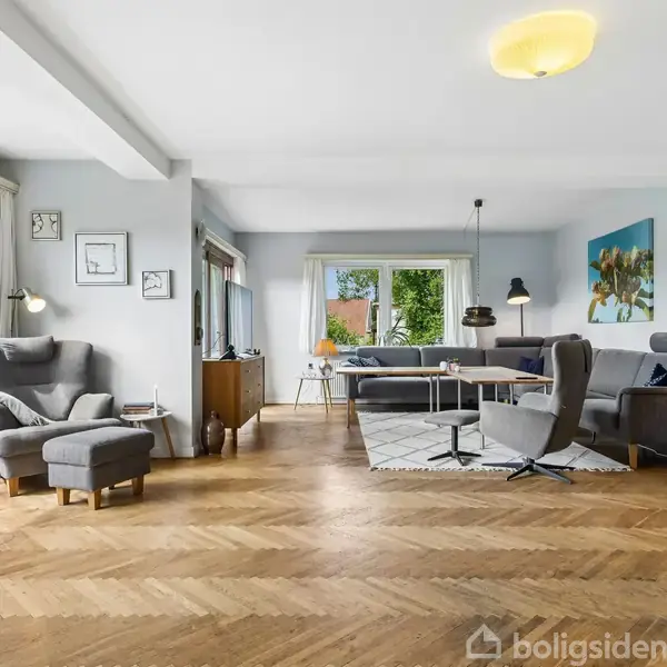 Stue med trægulv og lyseblå vægge, indrettet med sofaer og lænestol ved et sofabord. Store vinduer giver udsigt til grønt haveområde. Maleri og lamper hænger på væggene.